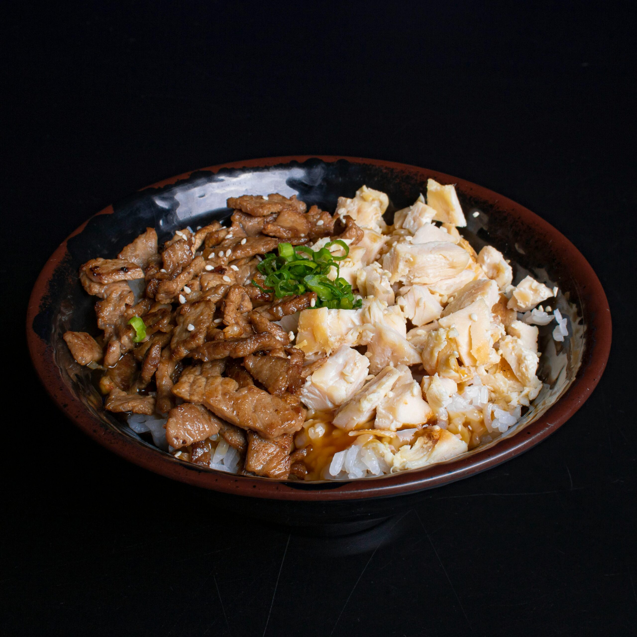 Close-up of a savory mixed meat rice bowl with garnish on a black background.