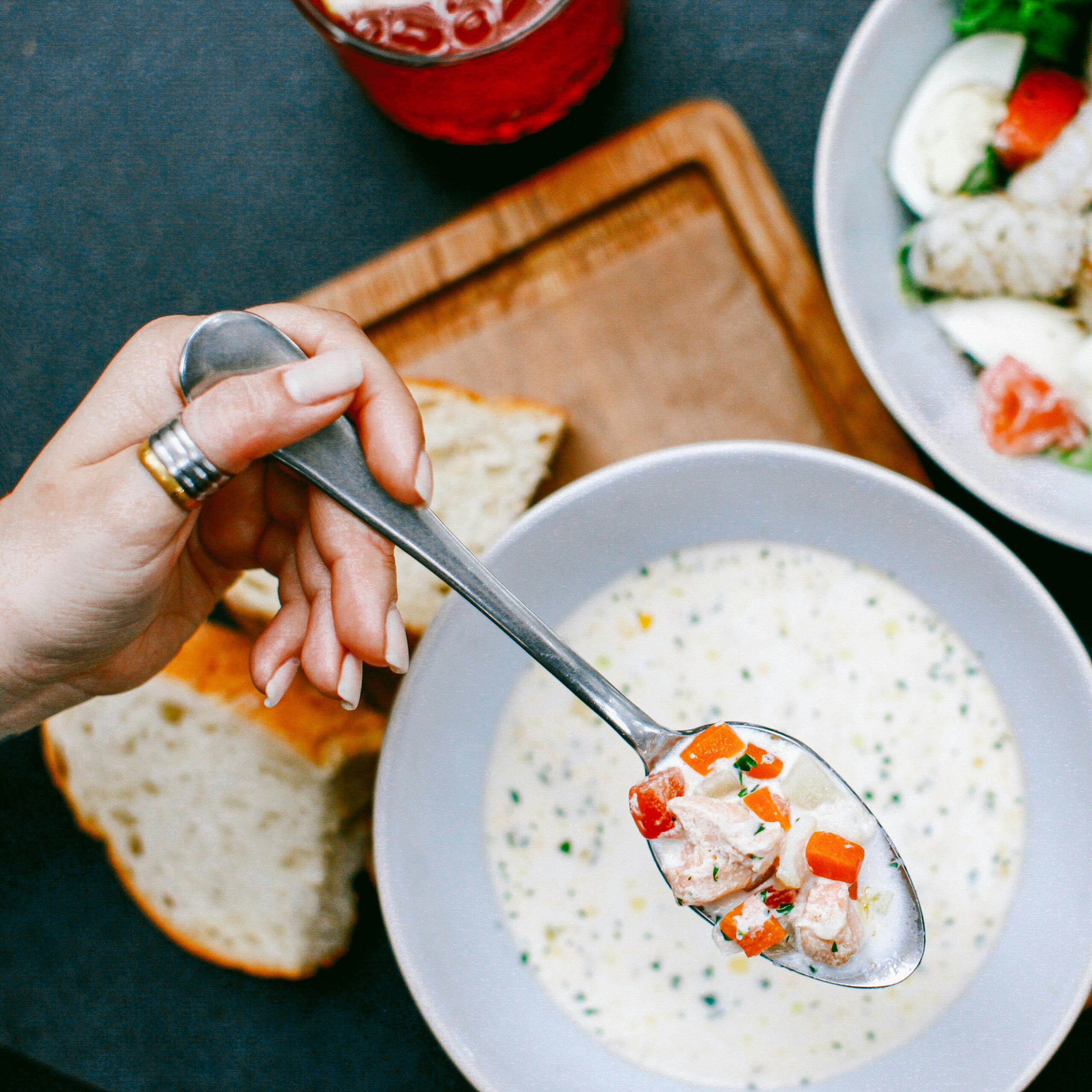 A close-up shot of a hand holding a spoonful of creamy soup with diced vegetables, accompanied by bread.