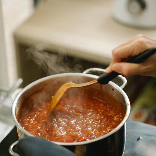 High angle crop faceless chef in potholder stirring bolognese sauce in pan stewing on stove in modern kitchen